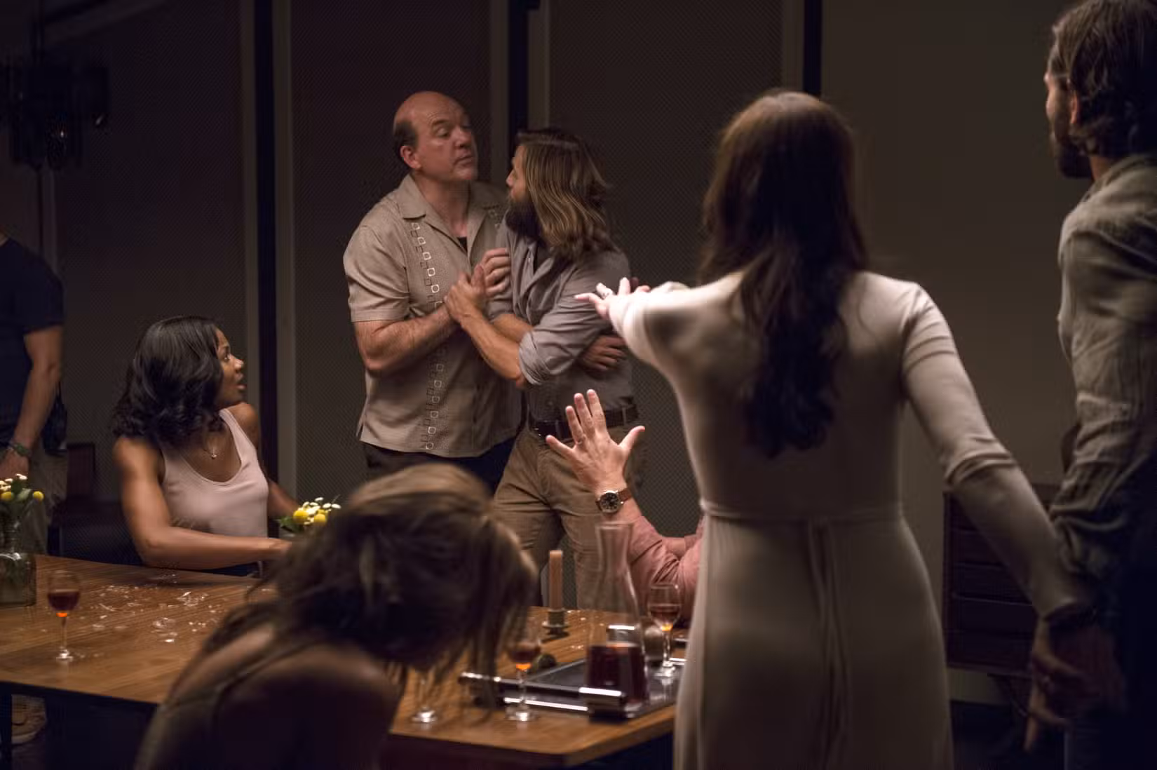 Em uma cena do filme O Convite, de 2016, um grupo de pessoas senta-se e fica parado ao redor de uma mesa de jantar parecendo controverso e confuso; um homem mais velho agarra uma mulher mais jovem enquanto outra mulher, de costas para a câmera, estende a mão como se tentasse impedi-los.