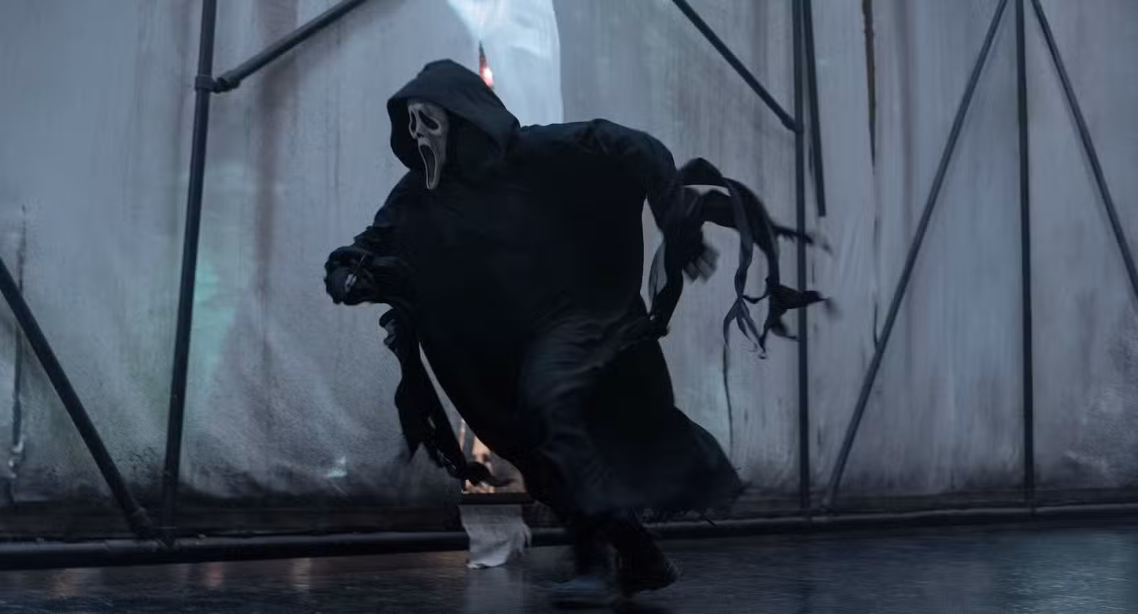 Ghostface, o assassino vestido de preto e máscara branca da franquia Pânico, corre atrás de alguém contra o pano de fundo de telas brancas sustentadas por andaimes pretos em Pânico VI