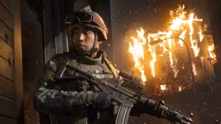 A soldier in Modern Warfare 4 looking behind him while holding an assault rifle with a blazing building burns in the background.