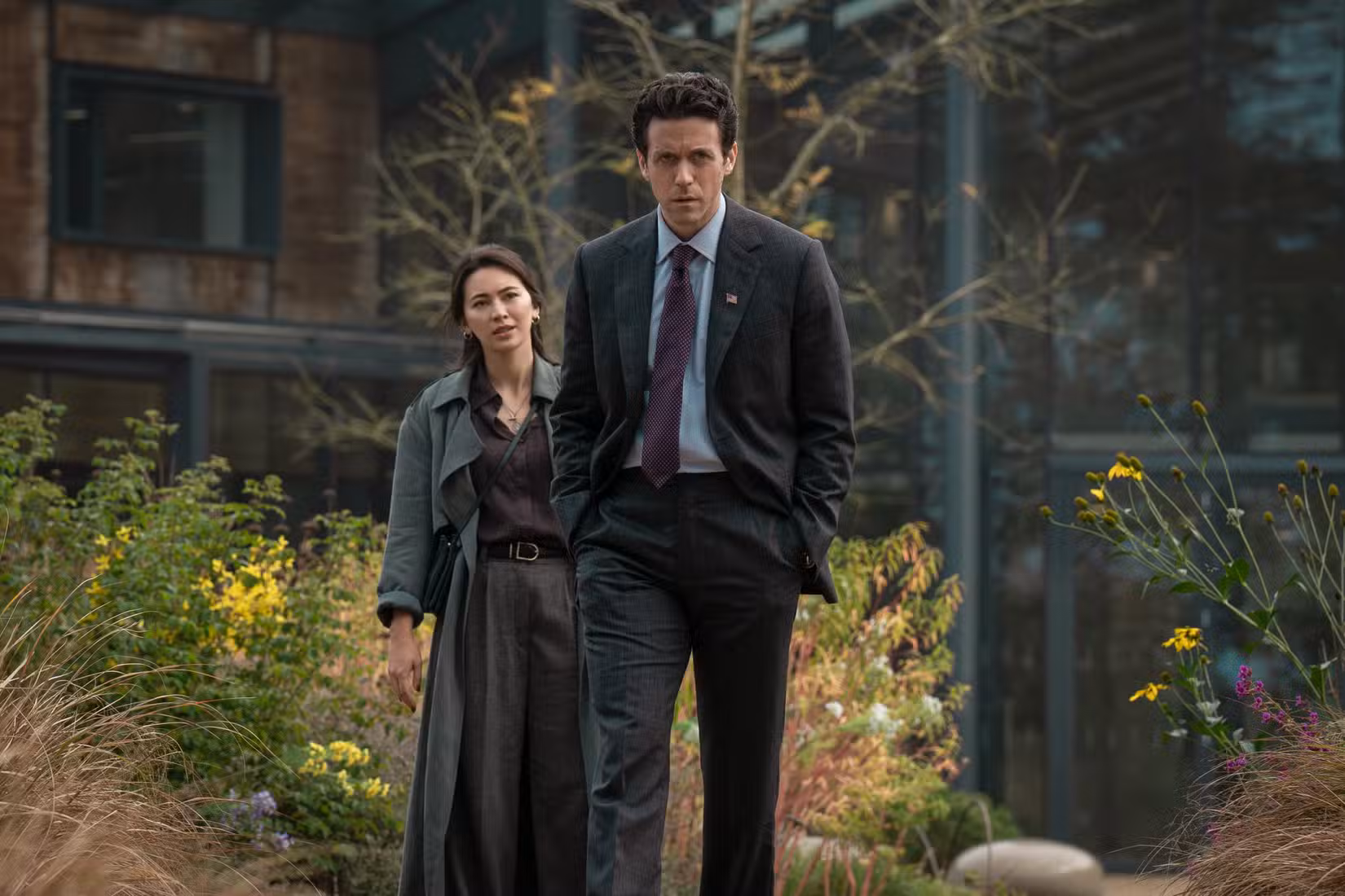 Daniel Keene (Ashley Zukerman) wearing a suit and tie walking in front of Helen Drew (Jessica Henwick) near some flowers in Silo season 3.